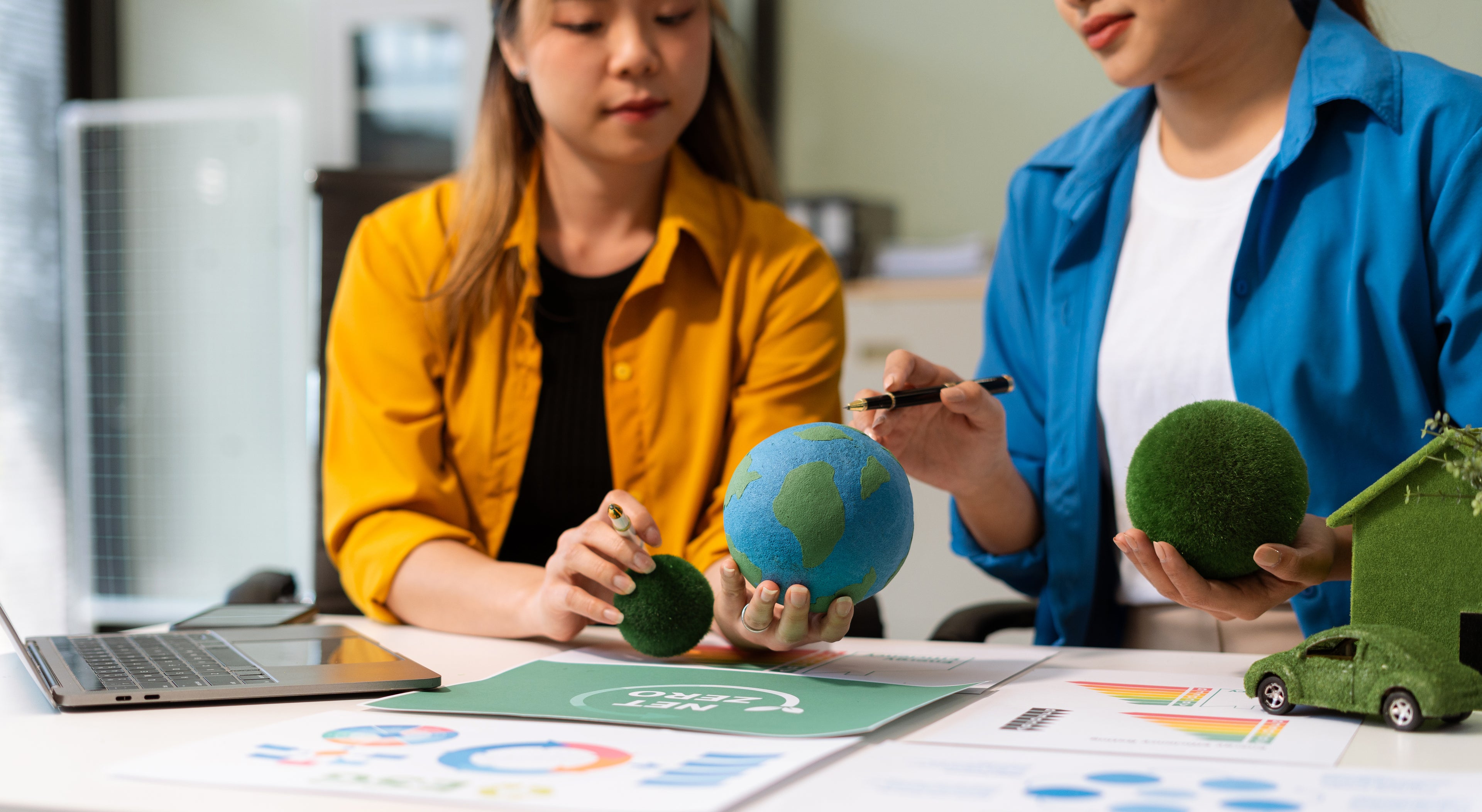 Twee collega’s overleggen over duurzaamheid en innovatie met symbolen zoals een wereldbol, huis en auto van gerecycled materiaal op de werktafel